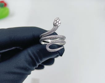 May include: A silver-toned metal ring shaped like a coiled snake. The ring features a textured surface that mimics snake scales, with a detailed head at one end and a tapered tail at the other. The metal has an oxidised finish to highlight the scales.