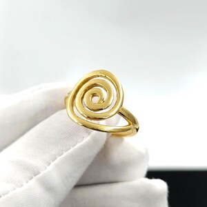 May include: A gold-colored ring featuring a spiral design. The ring's band is a simple, smooth design, and the spiral is the focal point. The ring is shown on a white glove, with a bright background.
