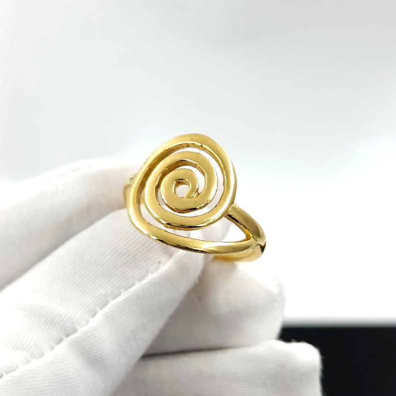 May include: A gold-colored ring featuring a spiral design. The ring's band is a simple, smooth design, and the spiral is the focal point. The ring is shown on a white glove, with a bright background.