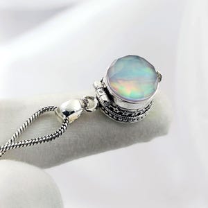 Puede incluir: Un collar con colgante de plata con una gran piedra preciosa facetada e iridiscente. La piedra preciosa está engastada en un engaste de plata detallado y ornamentado. La cadena es de plata delicada y texturizada. El colgante se muestra sobre un fondo blanco.