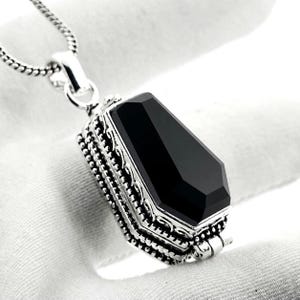May include: A silver pendant shaped like a coffin featuring a faceted black stone inlay on the top surface. The sides of the pendant are decorated with intricate metal scrollwork and beaded detailing. It hangs from a silver chain necklace.