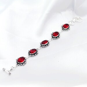 May include: A silver bracelet with five oval, deep red gemstones. Each gemstone is set in a decorative, beaded frame. The bracelet has a toggle clasp and is displayed on a white, textured surface.