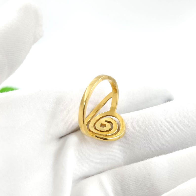 May include: A gold-colored ring with a spiral design. The ring features a band that curves up and around a central spiral, creating a unique and artistic look. The ring is set against a white background.
