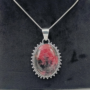 May include: A silver chain necklace with a pendant featuring a pink and black oval gemstone set in a silver frame with a beaded border.