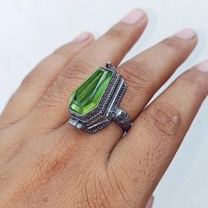 Op de afbeelding: Een zilverkleurige metalen ring met een doodskistvormig ontwerp en een langwerpige, gefacetteerde limoengroene steen in het midden. De metalen band heeft ingewikkelde, getextureerde details langs de zijkanten en randen van de zetting.