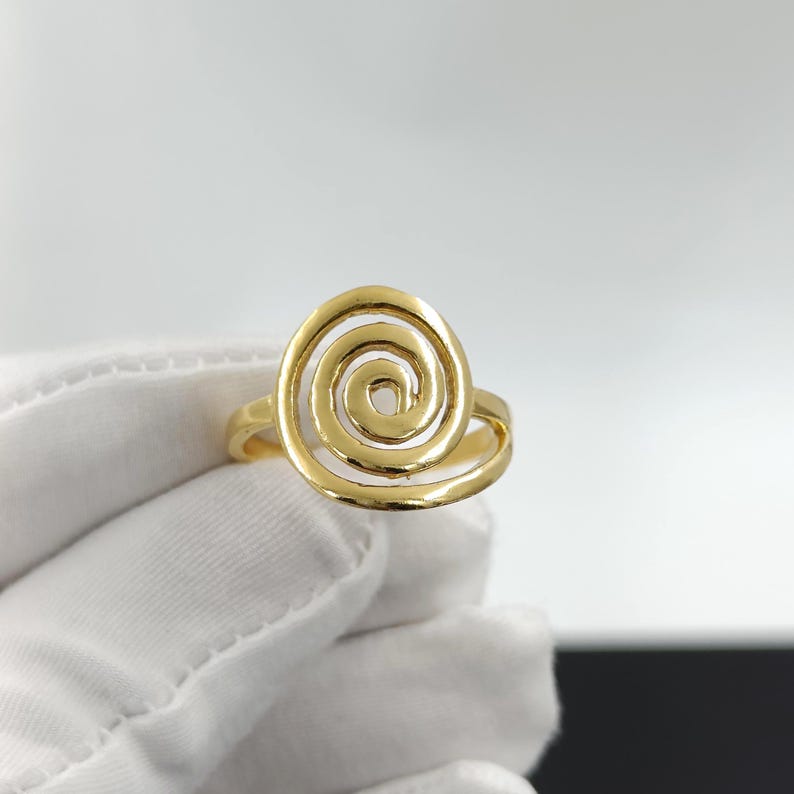 May include: A gold-colored ring featuring a spiral design. The ring's band is smooth, and the spiral is open, creating a unique and eye-catching piece of jewelry. The ring is shown against a white background.