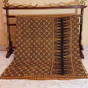 May include: A patterned textile in brown and black, displayed on a wooden rack with gold dragon ornaments. The textile has a diamond pattern and a central black panel with geometric designs. The rack features dragon head carvings.