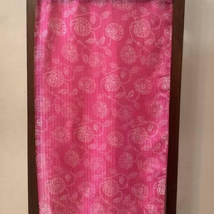 May include: A vibrant pink textile with a repeating floral pattern and a decorative border. The fabric is draped over a dark wooden frame with carved details at the top. The floral design is in a lighter shade of pink.