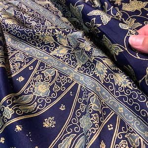 Puede incluir: Primer plano de una tela azul marino con un estampado floral y geométrico en tonos azul claro y dorado. La tela está doblada, revelando el intrincado diseño. El patrón incluye flores estilizadas, hojas y líneas onduladas, creando una apariencia rica y texturizada.
