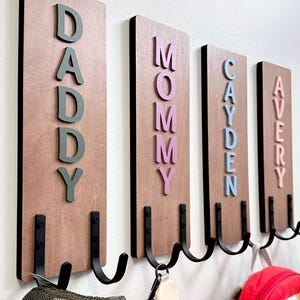 Könnte beinhalten: Wandgarderobe aus Holz mit vier vertikalen Tafeln. Jede Tafel trägt einen Namen in farbigen 3D-Buchstaben: DADDY, MOMMY, CAYDEN und AVERY. Schwarze Metallhaken sind darunter angebracht. Ein Hut, eine Handtasche und ein Rucksack hängen daran.