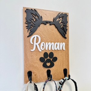 May include: Wooden dog leash holder with three hooks. The holder features a black silhouette of a dog's head with ears, a paw print with a heart, and the name "Roman" in white letters.