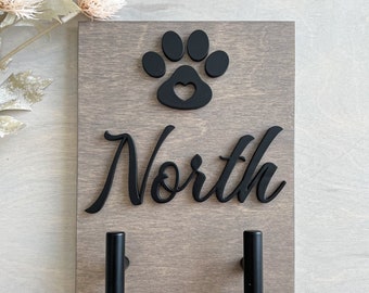 Puede incluir: Una placa de pared rectangular de madera con acabado gris que presenta una huella de pata negra en 3D con un recorte en forma de corazón, el nombre North en letras cursivas negras y dos ganchos cilíndricos negros en la parte inferior.