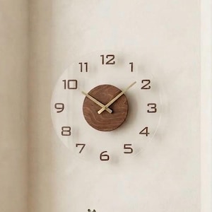 May include: A circular wall clock featuring a transparent acrylic face with dark brown numerals from one to twelve. The centre of the clock is a solid dark wood circle with gold-coloured hour, minute, and second hands mounted in the middle.
