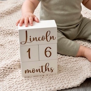 May include: A toddler wearing a sage green short-sleeved knit romper, holding a wooden rattle, sitting next to a stack of white wooden blocks. The blocks feature the name Lincoln, the number 16, and the word months in brown script lettering.