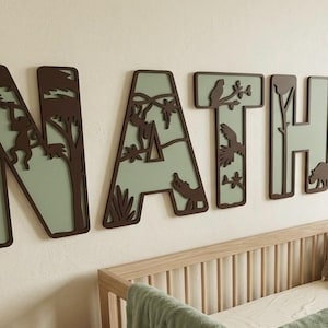 Op de afbeelding: Een kinderkamersc&egrave;ne met een houten wieg, een groene deken en knuffels. Boven de wieg staat de naam "NATHAN" in grote, decoratieve letters met een junglethema, inclusief dieren en bladeren.