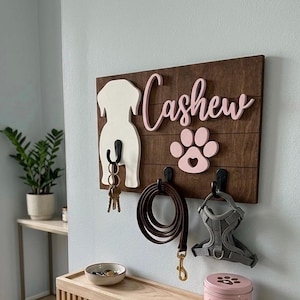 May include: A rectangular dark brown wooden wall plaque featuring a white dog silhouette, a pink paw print, and the name Cashew in pink script. Three black hooks hold a set of keys, a brown leash, and a gray harness. Below sits a light wood shelf.