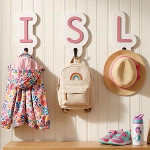 Könnte beinhalten: Vier weiße Wandhaken mit rosa Buchstaben, die ISLA bilden. Daran hängen eine geblümte Kapuzenjacke, ein beiger Rucksack mit Regenbogen-Aufnäher, ein Strohhut mit rosa Band sowie eine braune Lederleine mit einem Schlüsselbund.