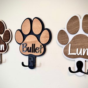 May include: Three wooden dog paw print hooks with black metal hooks. The paw prints are painted in brown and white with the names "Finn", "Bullet", and "Luna" written on them.