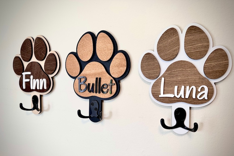 May include: Three wooden dog paw print hooks with black metal hooks. The paw prints are painted in brown and white with the names "Finn", "Bullet", and "Luna" written on them.