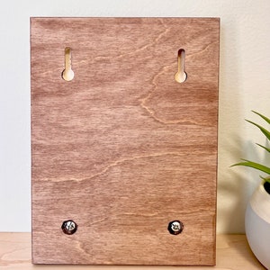 Puede incluir: La parte posterior de un letrero de madera con cuatro agujeros para tornillos para colgar. El letrero es de color marr&oacute;n claro.