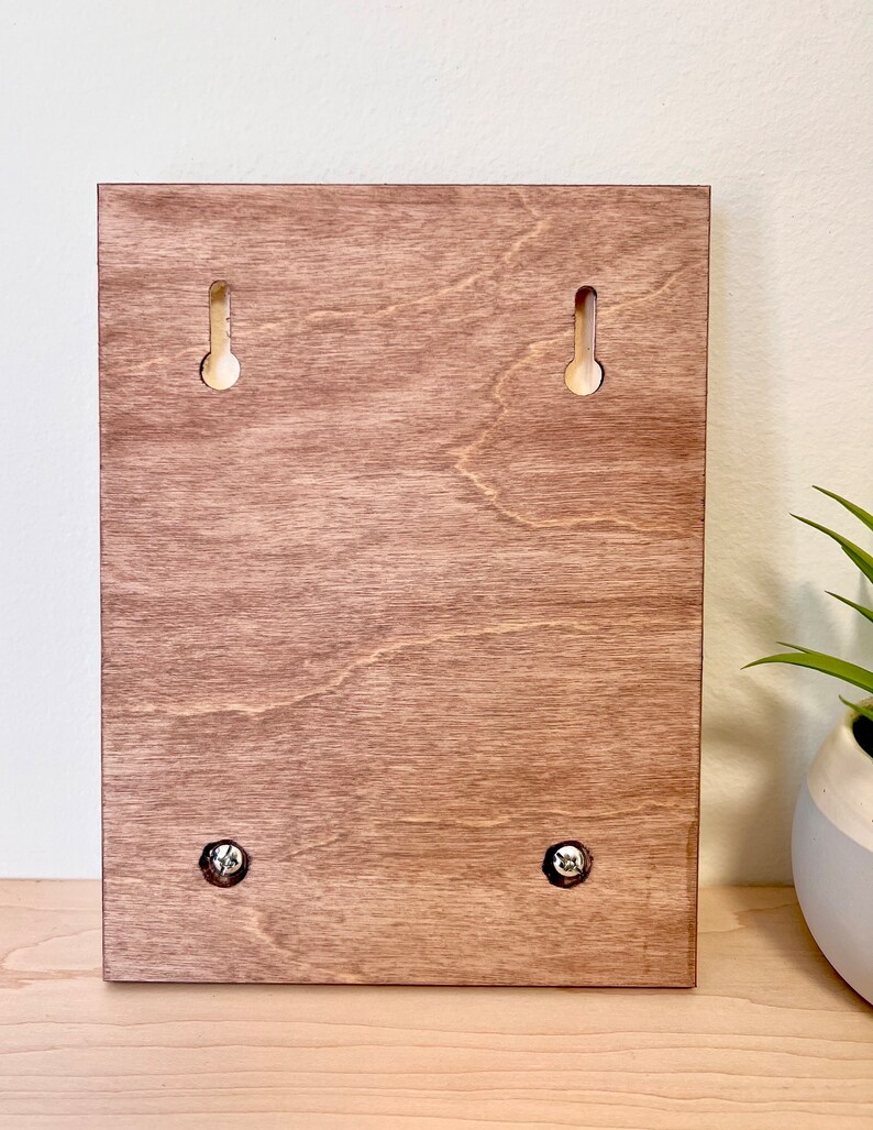 Puede incluir: La parte posterior de un letrero de madera con cuatro agujeros para tornillos para colgar. El letrero es de color marr&oacute;n claro.