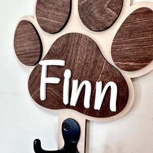 May include: Wooden dog paw print hook with the name "Finn" written in white letters. The hook is brown and has two black metal hooks.