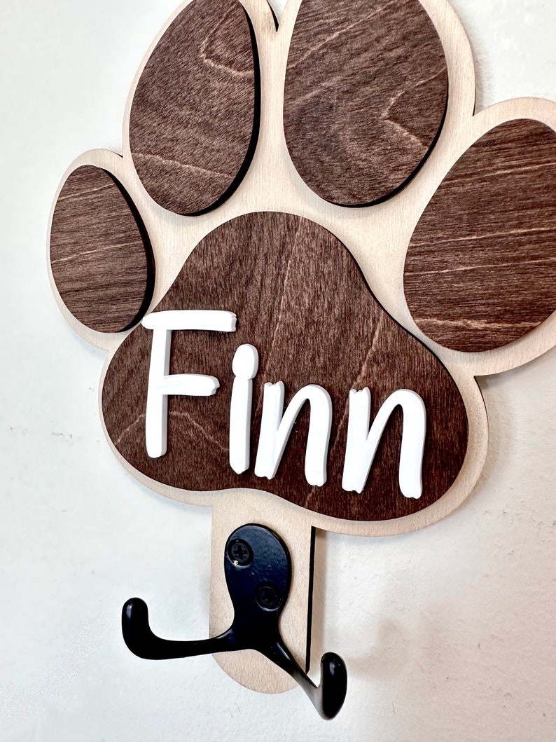 May include: Wooden dog paw print hook with the name "Finn" written in white letters. The hook is brown and has two black metal hooks.