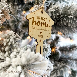 May include: A wooden key-shaped Christmas ornament with the text "OUR FIRST home", address, and the year 2022. The ornament is hanging on a snow-covered Christmas tree. The ornament has a house-shaped top and a key-shaped bottom.