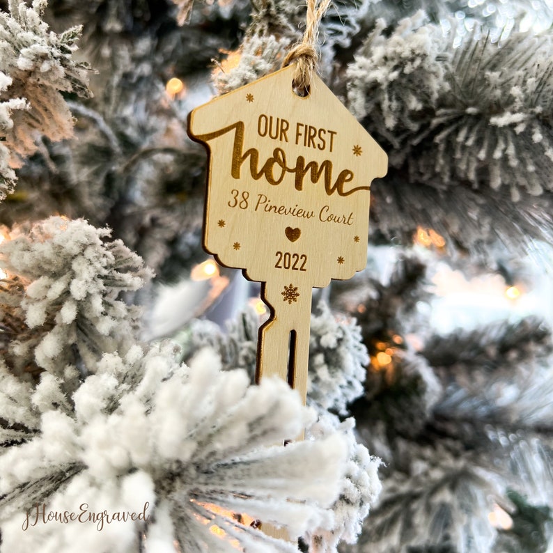 May include: A wooden key-shaped Christmas ornament with the text "OUR FIRST home", address, and the year 2022. The ornament is hanging on a snow-covered Christmas tree. The ornament has a house-shaped top and a key-shaped bottom.