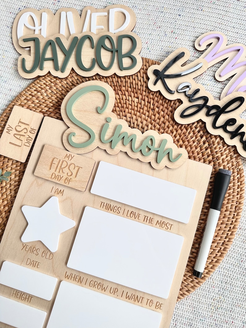 May include: A wooden milestone board featuring a light green cutout name Simon, white acrylic sections for writing, and engraved text including My First Day Of, Things I Love The Most, I Am, Years Old, Date, Height, and When I Grow Up, I Want To Be.