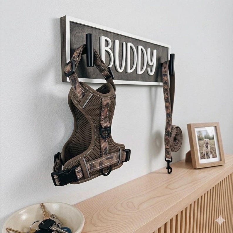 May include: A dog harness and leash set in a camouflage pattern hang from a wall-mounted sign that reads "BUDDY" in white letters. A small framed photo of a dog sits on a wooden shelf, with a bowl of keys in the foreground.