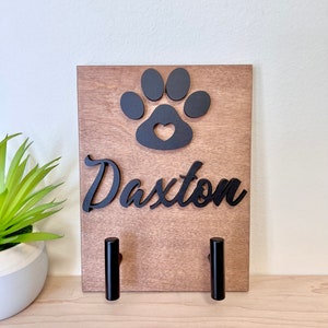 Puede incluir: Porta-correas de perro de madera con una huella de pata negra y un dise&ntilde;o de coraz&oacute;n. El porta-correas tiene dos ganchos negros y el nombre "Daxter" est&aacute; escrito en letras cursivas negras.