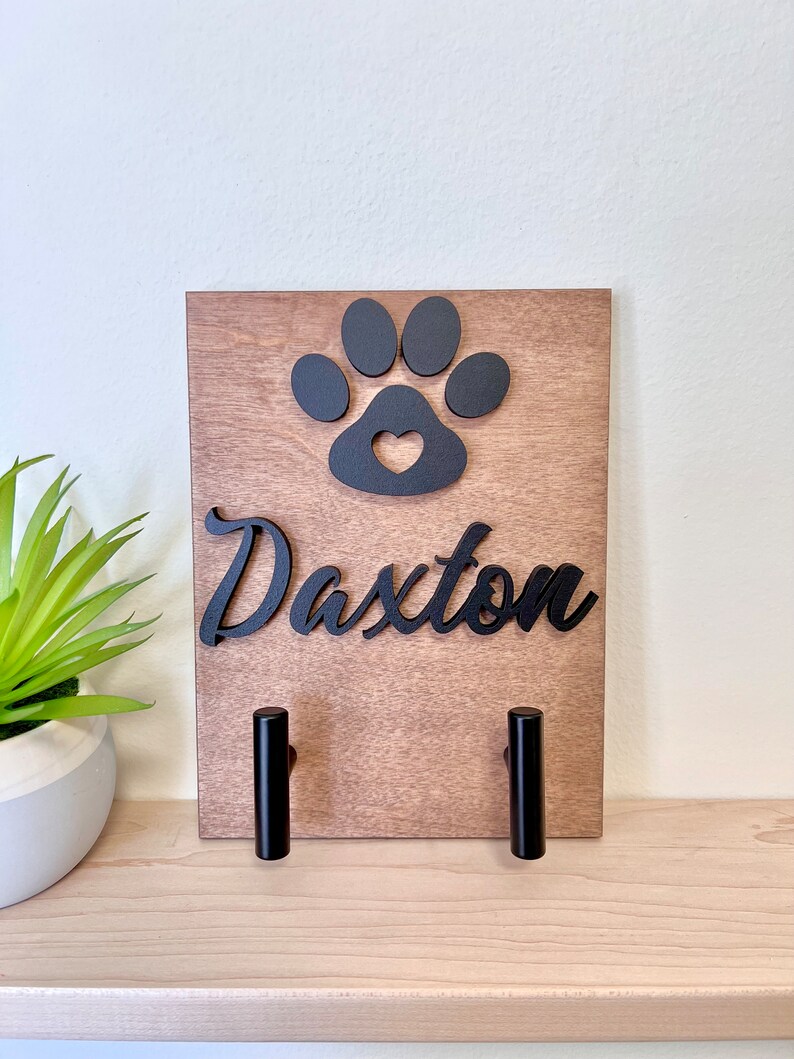 Puede incluir: Porta-correas de perro de madera con una huella de pata negra y un dise&ntilde;o de coraz&oacute;n. El porta-correas tiene dos ganchos negros y el nombre "Daxter" est&aacute; escrito en letras cursivas negras.