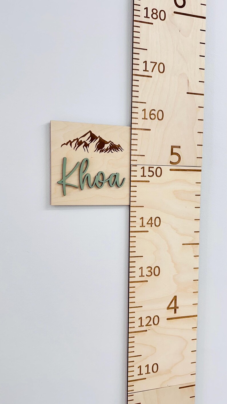 Puede incluir: Un medidor de crecimiento de madera con un dise&ntilde;o de monta&ntilde;a y el nombre "Khoa" en letras verdes. El medidor mide de 110 a 180 pulgadas.