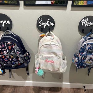 Puede incluir: Tres placas de pared redondas de madera negra con los nombres Logan, Sophia y Mason en letra blanca, cada una con dos ganchos de metal negro. Tres mochilas cuelgan de estos ganchos: una mochila azul marino de b&eacute;isbol, una mochila blanca iridiscente y una mochila azul estampada.