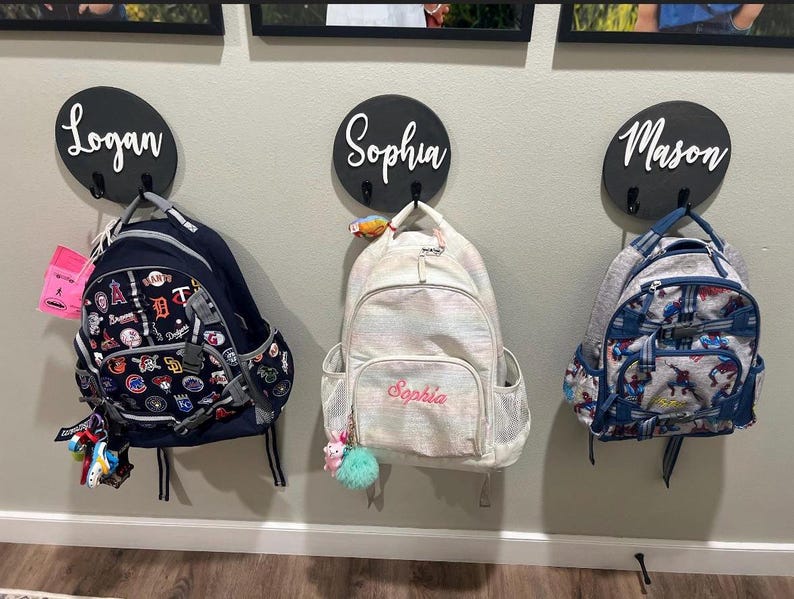 Puede incluir: Tres placas de pared redondas de madera negra con los nombres Logan, Sophia y Mason en letra blanca, cada una con dos ganchos de metal negro. Tres mochilas cuelgan de estos ganchos: una mochila azul marino de b&eacute;isbol, una mochila blanca iridiscente y una mochila azul estampada.