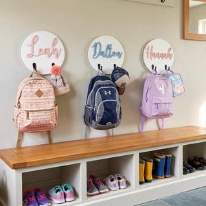 Puede incluir: Tres placas circulares blancas montadas en la pared con los nombres Leah, Dalton y Hannah en letras cursivas en relieve. Cada placa tiene dos ganchos negros que sostienen mochilas, un gorro y una bolsa de almuerzo. Debajo hay un banco de madera blanco.