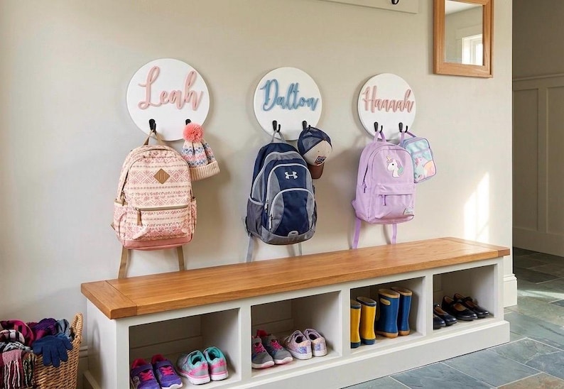 Puede incluir: Tres placas circulares blancas montadas en la pared con los nombres Leah, Dalton y Hannah en letras cursivas en relieve. Cada placa tiene dos ganchos negros que sostienen mochilas, un gorro y una bolsa de almuerzo. Debajo hay un banco de madera blanco.