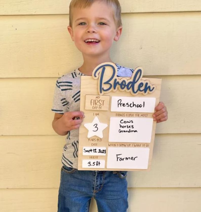 May include: A rectangular wooden milestone board featuring the name Broden in blue script. The board includes white sections for writing details like preschool, age 3, date Sept 12 2023, height 3.5 ft, favorite things including cows, horses, and Grandma, and future career as a farmer.