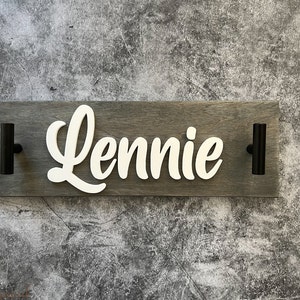 May include: A wooden sign with the name "Lennie" in white cursive lettering. The sign is gray and has two black handles. The background is a textured gray surface.