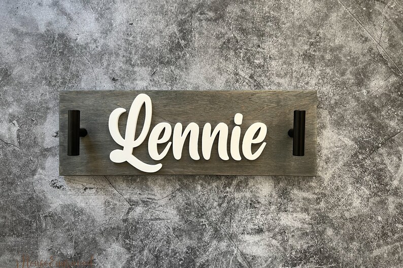 May include: A wooden sign with the name "Lennie" in white cursive lettering. The sign is gray and has two black handles. The background is a textured gray surface.