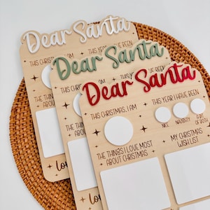 May include: Three wooden Dear Santa boards with white, green, and red raised lettering. Each board features text including This Christmas I am, This Year I Have Been, and My Christmas Wish List, with blank white spaces for writing and star accents.