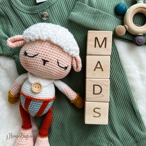 May include: A handmade crochet lamb doll with a white head, pink ears, and closed eyes. The doll is wearing a white shirt with a wooden button and a red, gray, and orange outfit. Wooden blocks spell out the name "MADS".