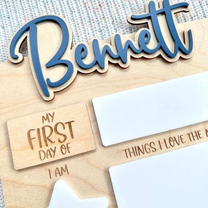 May include: A light-colored wooden milestone board featuring the name Bennett in raised blue script lettering. The board includes engraved text reading MY FIRST DAY OF, I AM, and THINGS I LOVE THE MOST, alongside blank white rectangular and star-shaped areas.