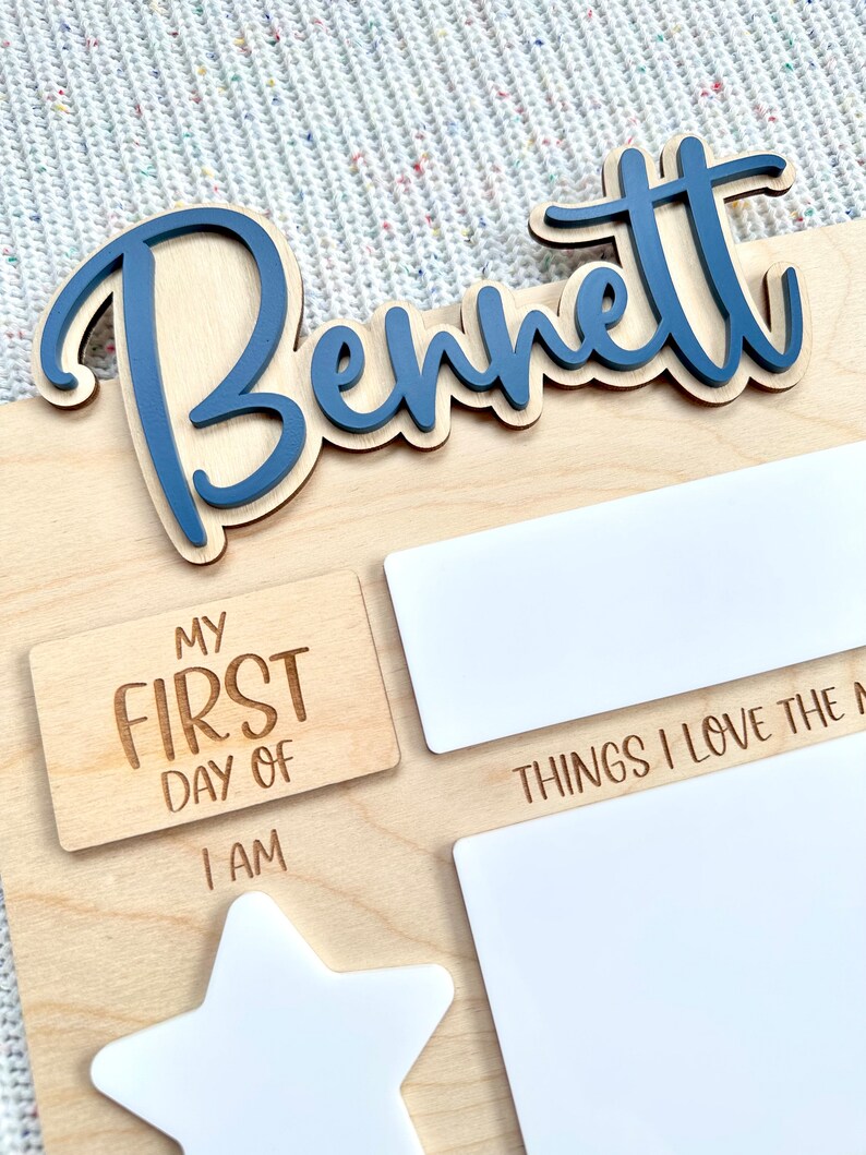 May include: A light-colored wooden milestone board featuring the name Bennett in raised blue script lettering. The board includes engraved text reading MY FIRST DAY OF, I AM, and THINGS I LOVE THE MOST, alongside blank white rectangular and star-shaped areas.