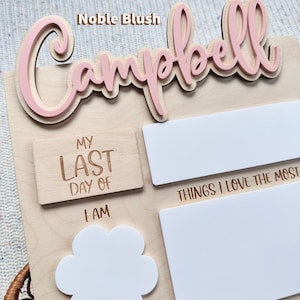 May include: A light-colored wooden milestone board featuring the name Campbell in raised pink script. The board includes engraved text such as My Last Day Of, I Am, Years Old, and Things I Love The Most, with blank white spaces for writing.