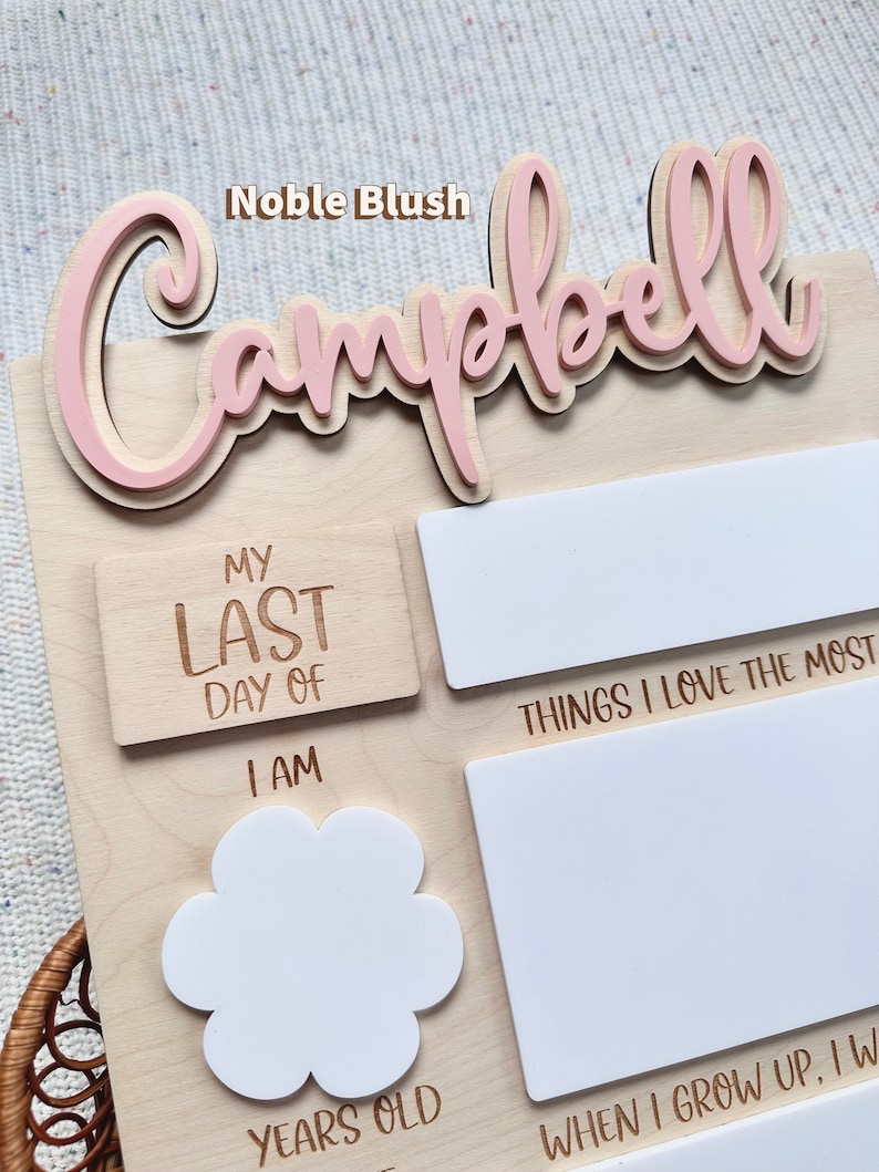 May include: A light-colored wooden milestone board featuring the name Campbell in raised pink script. The board includes engraved text such as My Last Day Of, I Am, Years Old, and Things I Love The Most, with blank white spaces for writing.