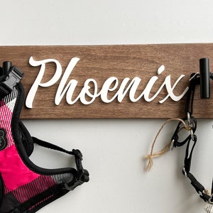 May include: A wooden dog leash holder with the name "Phoenix" in white script. Two black hooks hold dog harnesses, one pink and black plaid, the other black and tan. A pet accessory for dog owners.