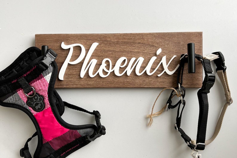 May include: A wooden dog leash holder with the name "Phoenix" in white script. Two black hooks hold dog harnesses, one pink and black plaid, the other black and tan. A pet accessory for dog owners.
