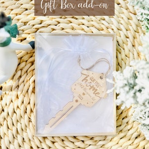 May include: A wooden key-shaped ornament with the text "OUR FIRST HOME" and the address "1942 Sturgeon Rd 2023" inside a clear gift box. The gift box is inside a woven basket. The words "Gift Box add-on" are at the top.
